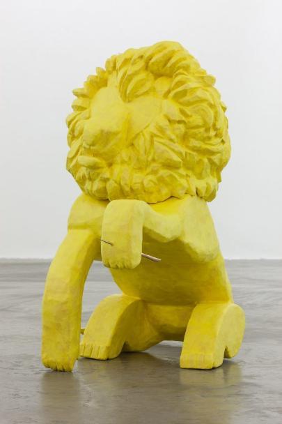 photo of a sculpture of a yellow lion sitting on the grey floor of a gallery. The lion sits on his back legs, with his front left paw extended upwards towards the camera. The left paw has a large wooden thorn through it. The lion is painted a cheerful bright yellow all over.