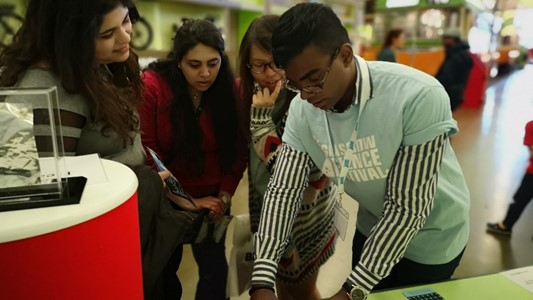 photo of a member of the CSPE team (a black young man) demonstrates illusions to three onlookers at the Glasgow Science Festival