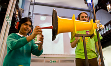 Photograph showing a child and a demonstrator both wearing ear defenders, the child is blowing an air horn into a cone shaped object.