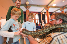Photograph of a man with a large snake around his neck, two children are touching the snake whilst an adult oversees. 