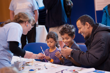 Photograph showing a man with two children, a GSF volunteer is handing out colouring pencils. 