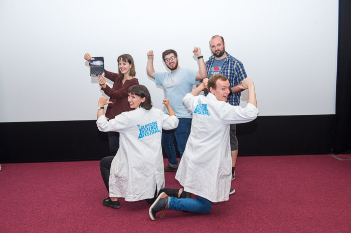 Photograph showing 5 people, 2 are wearing GSF lab coats and 1 is holding up a poster for 'Chasing the Waves' stage show. 
