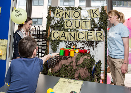 Photograph of a child throwing plastic balls at colourful cans under the sign 'Knock out Cancer'.