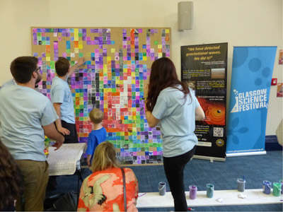 Photograph showing three GSF volunteers with a colourful painted mural and posters about the detection of gravitational waves. 