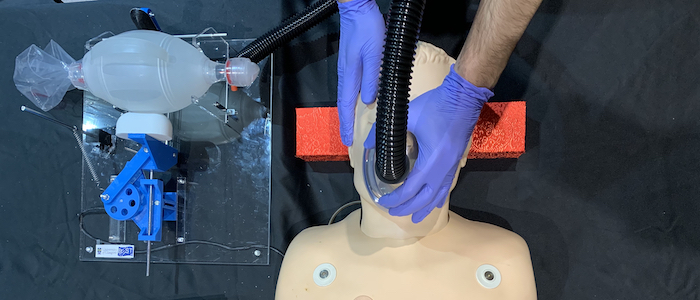 A pair of gloved hands hold a mask over the face of a medical dummy during a test of the GlasVent system. The GlasVent system sits beside the dummy, connected to the mask by a length of plastic tubing.