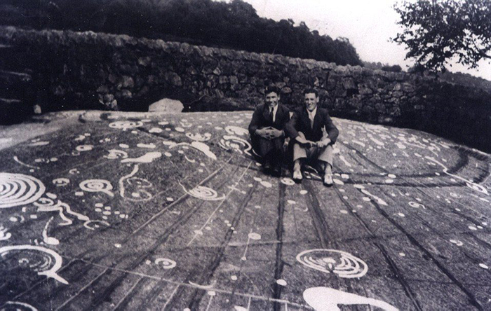 A black and white image of two young men sitting on the Cochno Stone before its burial in 1965. White markings were painted onto the stone by Mann in 1937.