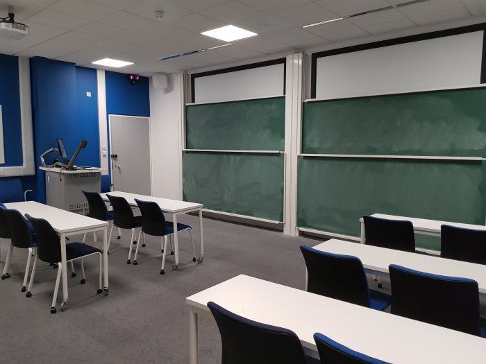 Flat floored teaching room with tables and chairs in rows, chalkboards, screens, projector, visualiser, and PC