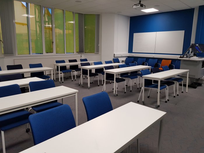 Flat floored teaching room with rows of tables and chairs in rows, whiteboards, visualiser, projector, PC, and lectern.