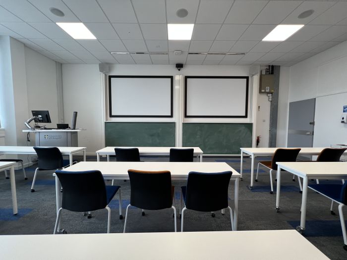 Flat floored teaching room with tables and chairs in rows, chalkboards, screens, visualiser, and PC