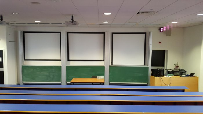 Raked lecture theatre with fixed seating, blackboards, screens, projectors, and PC