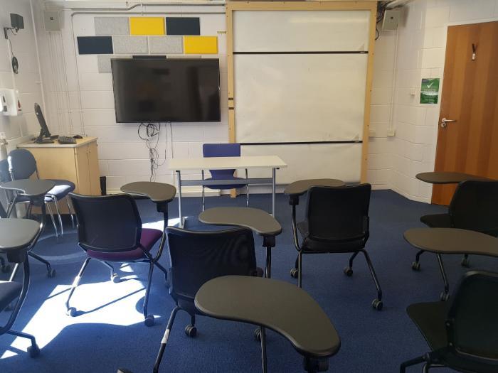 Flat floored teaching room with tablet chairs, whiteboard, large monitor, lecturer's table and chair, and PC.