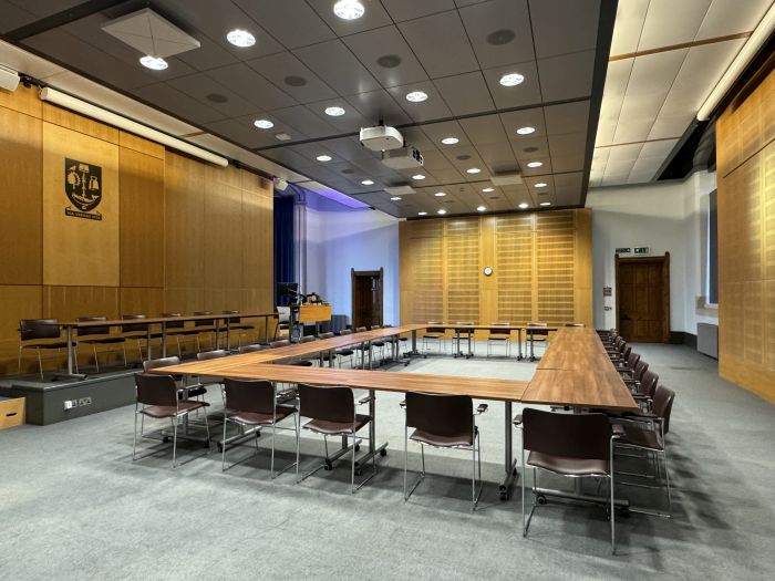 Flat floored room with tables and chairs in a boardroom layout, raised podium with tables and chairs in a row, projector, lectern, and PC.