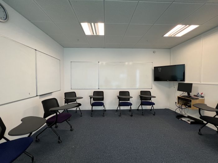 Flat floored teaching room with tables and chairs in round table set-up, whiteboard, video monitor, and PC