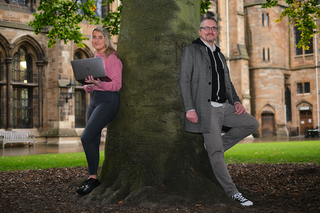 Graduate Apprentice in Software Development Megan Gallagher, holding a laptop, leans against a tree in the University quad while Dr Matthew Barr of the School of Computing Science leans on the other side