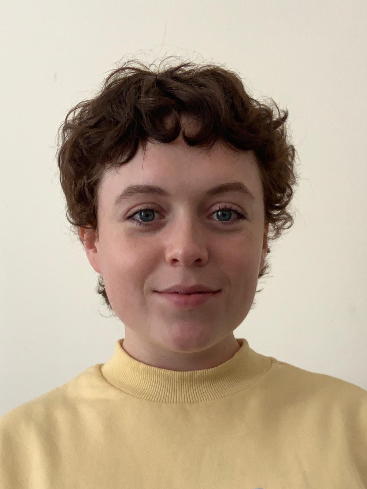 A young white woman with short brown hair wearing a light yellow jumper, smiling. 