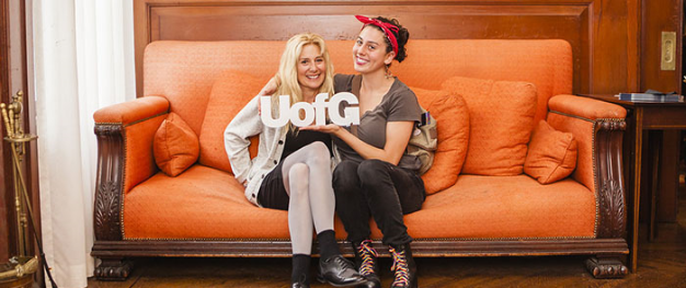Two women sitting on a sofa with a small UofG sign