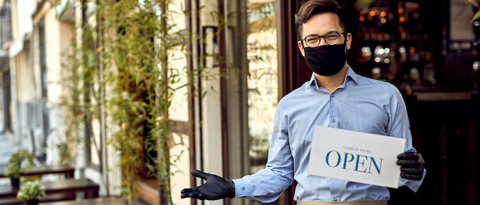 A masked and gloved waiter stands outside a restaurant, holding a sign that reads 'OPEN'