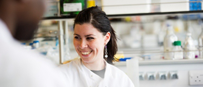 Woman in lab coat smiling at someone she is talking to who has their back to the camera