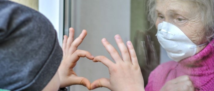 Child forming a heart shape with hands at window with elderly woman watching