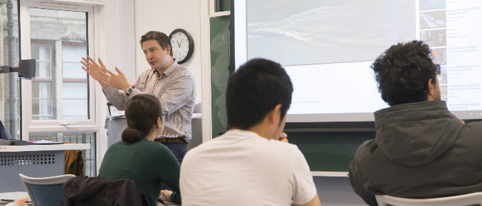 An individual standing at the front of a group sat at desks providing training