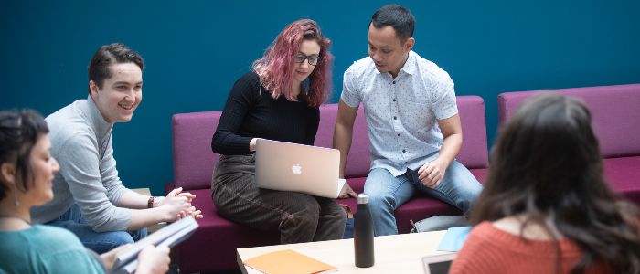 A group collaborating sat around a table with one individual taking the lead by showing a colleague information on a laptop