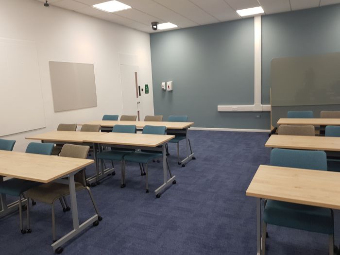 Flat floored teaching room with rows of tables and chairs, and movable glassboard. 