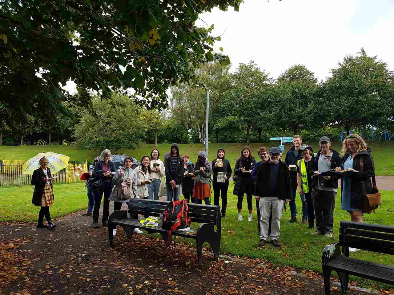 A group of people is in a park. The group is made of about 20 people who are standing up in front of two dark green benches. 