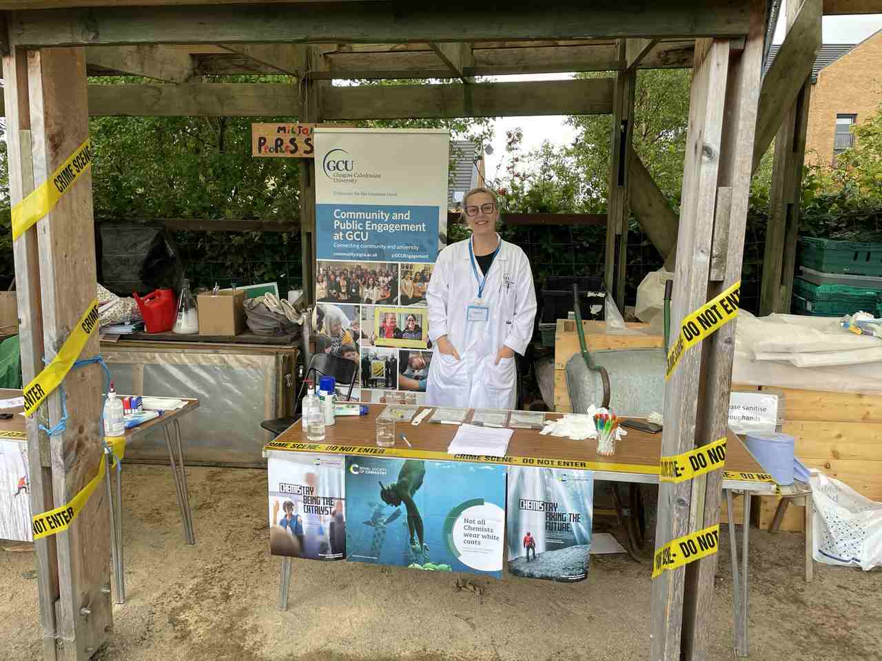 A wooden gazebo has yellow ‘crime scene – do not enter’ tape wrapped around the front posts. Under the gazebo from left to right are gardening items, a Glasgow Caledonian University sign, a female adult in a white lab coat and a wheelbarrow. In front of this is a table with worksheets and pens with posters advertising about chemistry hung from the front of the table. 