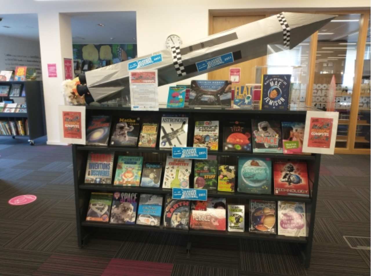 A bookshelf containing books about science with a focus on space. Above the bookshelf is a cardboard 3D model of a rocket which says ‘Glasgow Science Festival 2021’ on the side.