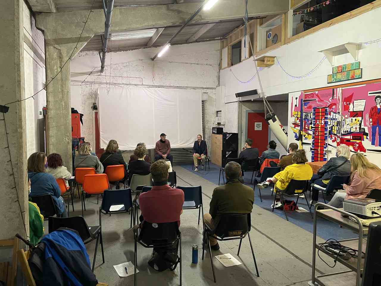 A group of people is indoors, sitting in front of two adults, who are also sitting down. Behind the two adults, there is a white projection screen. The two adults are speaking and moving. The audience in front of them is still and listening to them talk. 