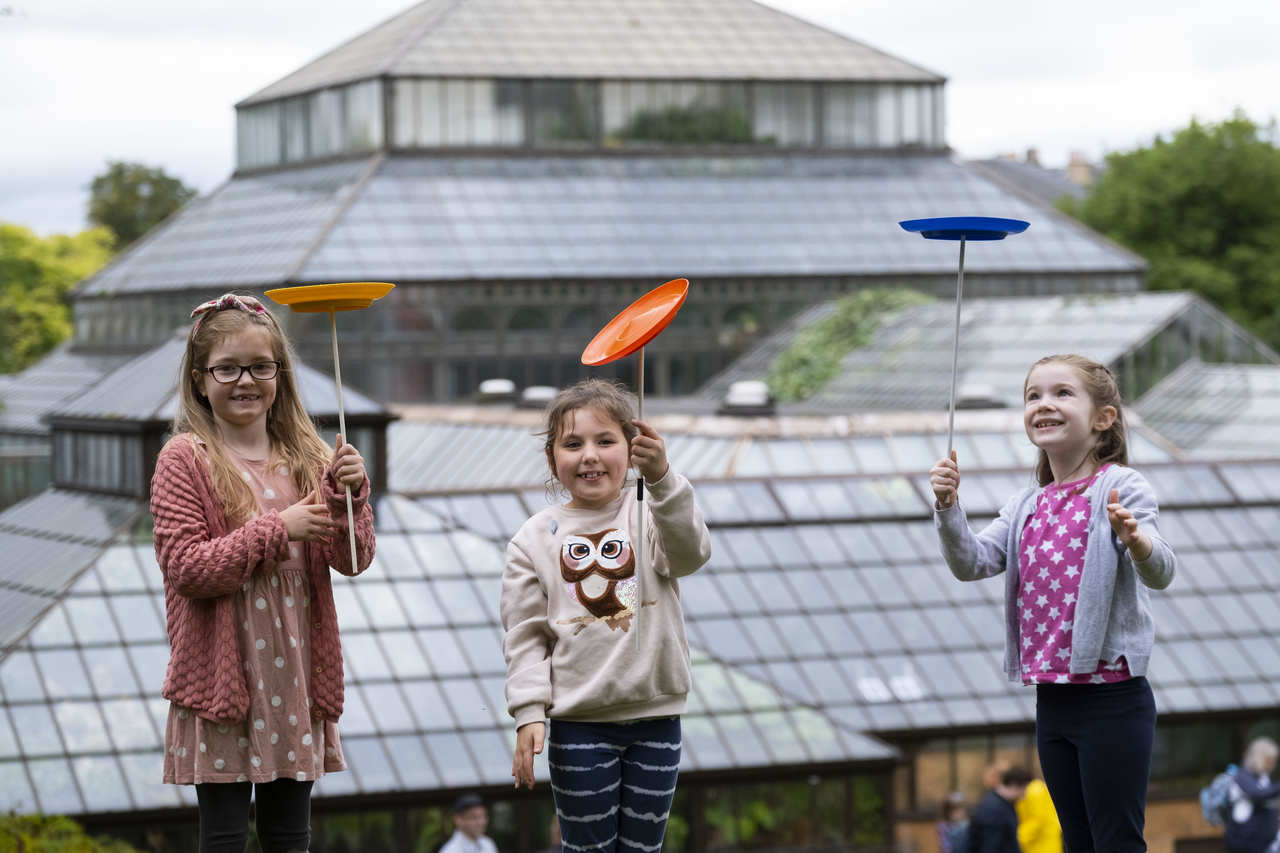 Three young girls are outside practicing their circus skills by balancing a plate on a stick and spinning it around. 