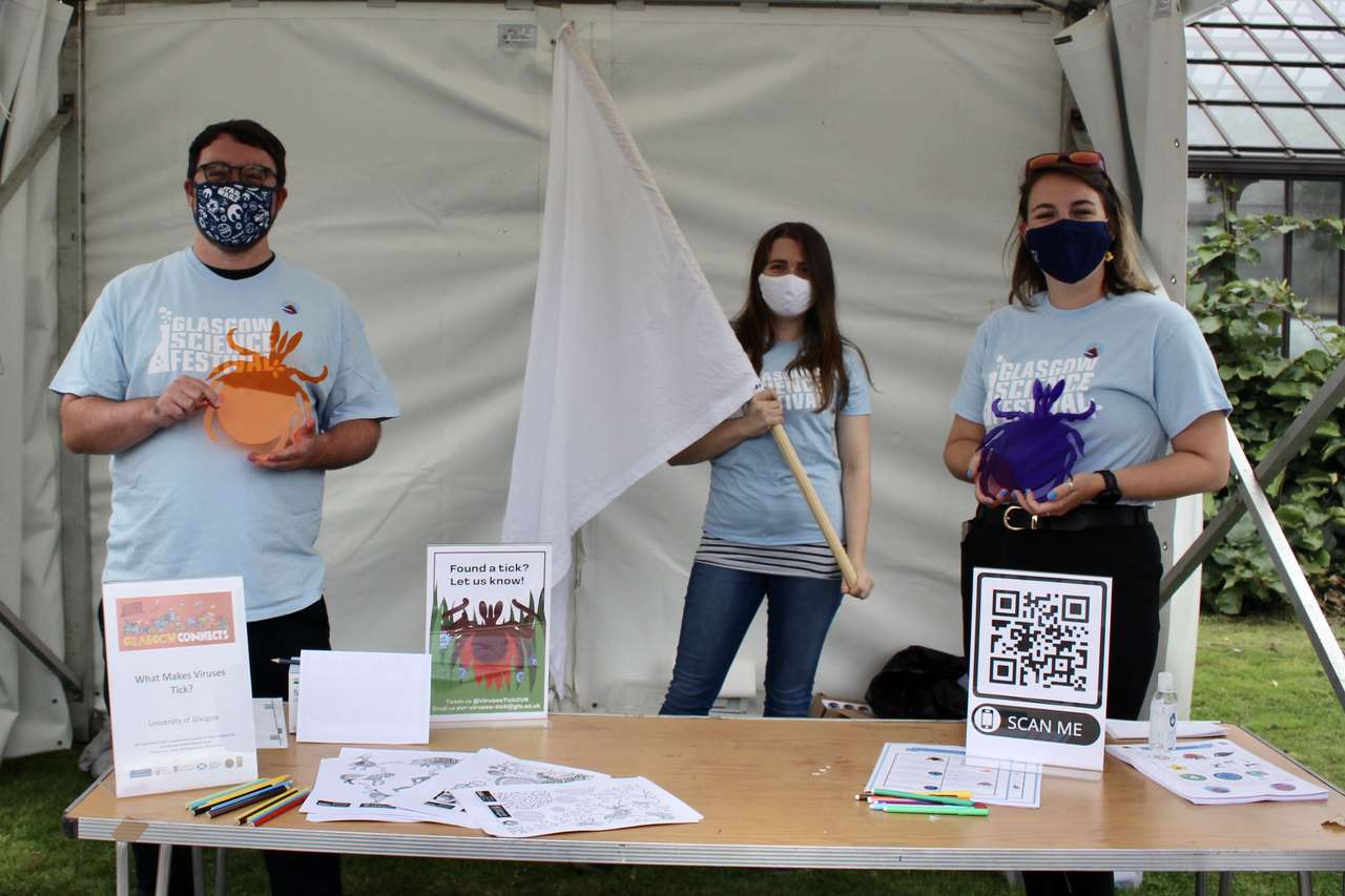 From left to right, there is an adult male wearing a mask and holding a giant plastic tic, a female with a large white flag and then another female holding a giant plastic tic. The table in front of them shows more information about tics. 