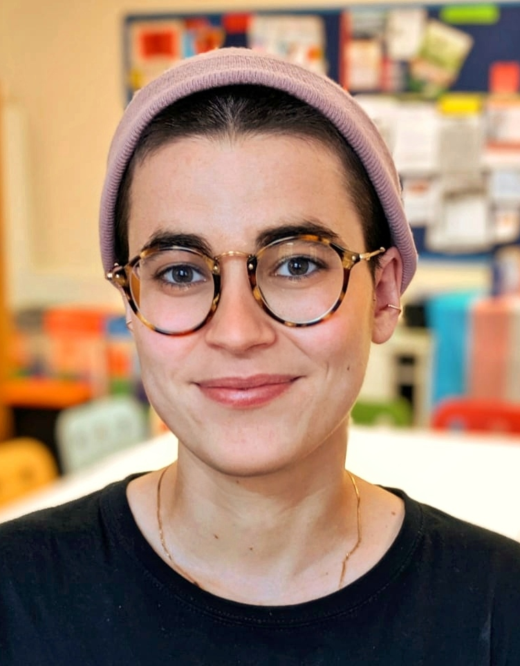 A headshot of a researcher smiling to the camera. She is white, with short brown hair, she has glasses and is wearing a black t-shirt and a pink beanie. 