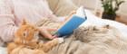 Photo of a woman reading a book with a fluffy ginger cat beside her