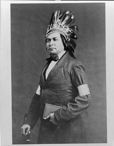 A man in formal standing pose wearing a long dress coat, and a feather headress