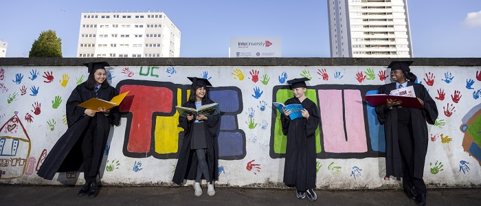 Four students wearing graduation gowns stand by a brightly coloured wall with a sign saying The Hub. They children are involved in the launch of IntoUniversity Maryhill centre 