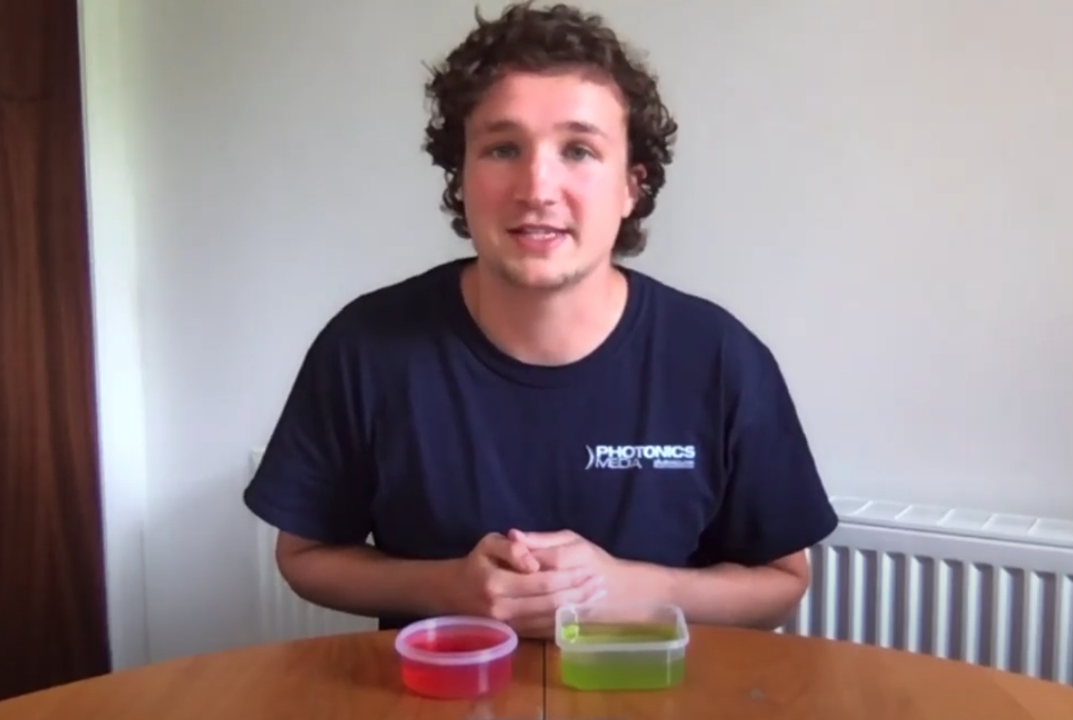 Man in blue shirt sits at desk with two tubs of different coloured jelly for online demonstration video. 