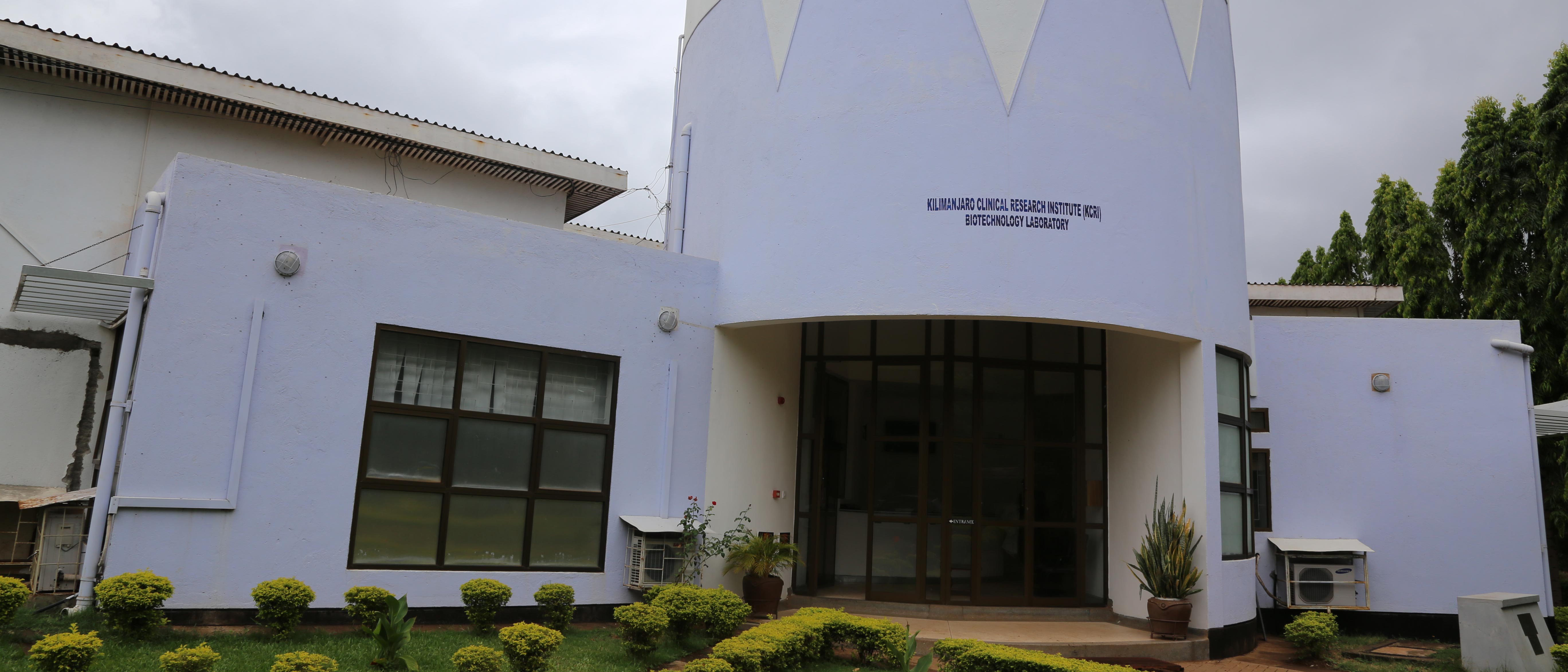 Outside view of the Kilimanjaro Clinical Research Institute, a light blue and white building with grass and plants in front.