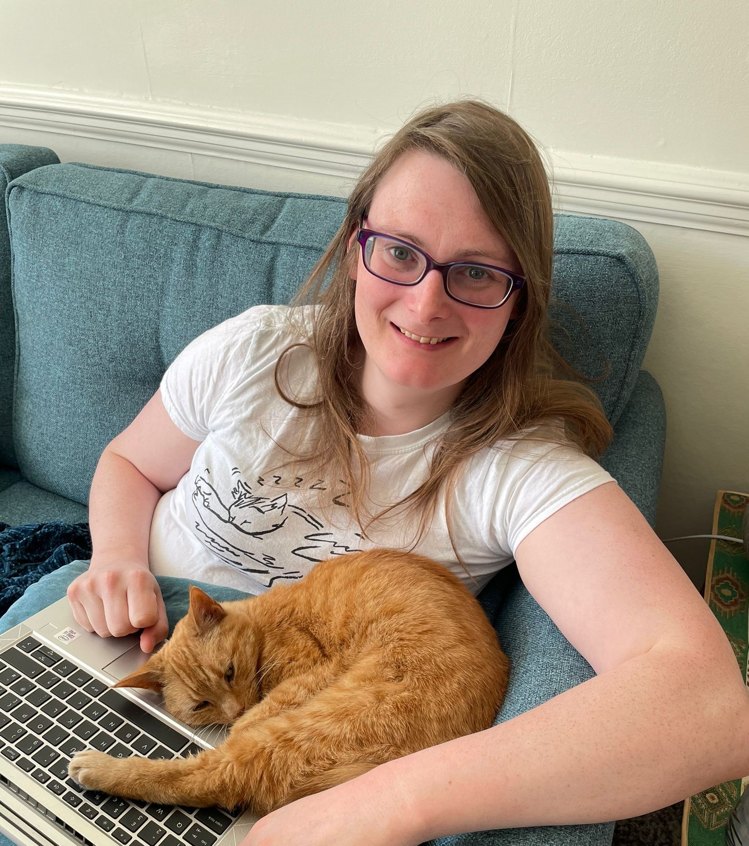 A smiling white woman sitting on a chair with a laptop, and a cat curled up into her.