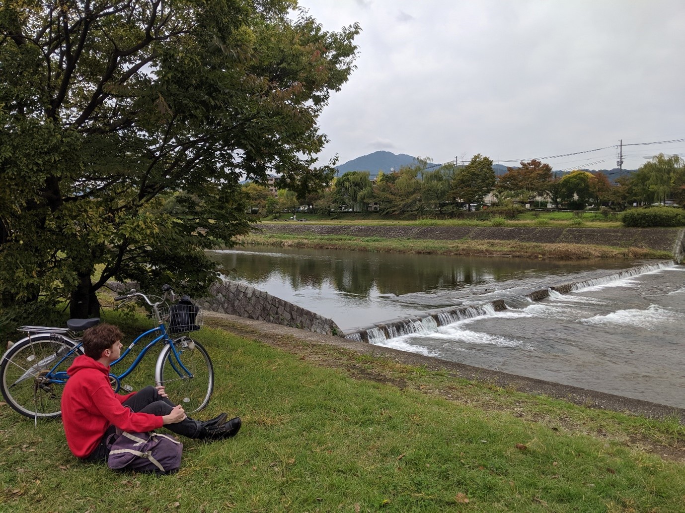 Calum Bell sitting by Kamo river near Kyoto University with Japanese bike