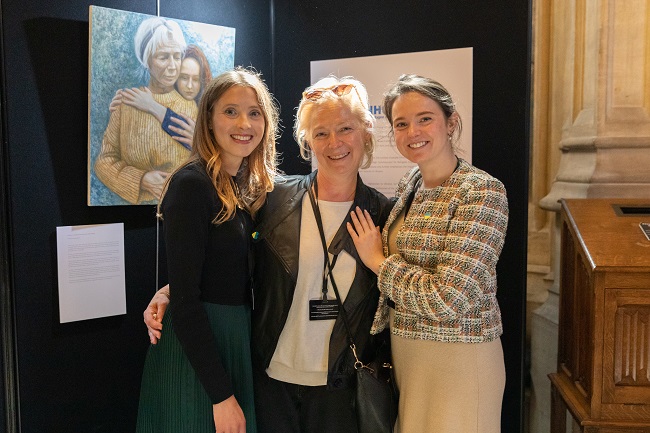 A photo of Hannah Thomas with Maria and her mother Nadia (in the middle) with Hannah's painting of them in the background