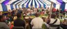 A large group of people sit and stand in a large circle under a blue and white striped tent. They are playing instruments and singing.