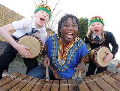 A black man plays a marimba and 2 laughing white children play drums