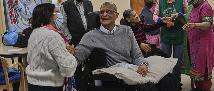 In a community room, an older Asian man in a wheelchair holding hands and smiling at a woman. There are other people chatting in the background. Source: Centre for Aging Better https://ageingbetter.resourcespace.com/pages/view.php?ref=11876&k=&search=age%2C+positive%2C+image%2C+library%2C+%40%40339&offset=0&order_by=resourceid&sort=DESC&archive=