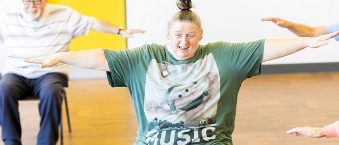 A happy woman taking part in an exercise class, sitting on chair and with her arms outstretched. There is an older man on a chair behind herSource: Centre for Aging Better
