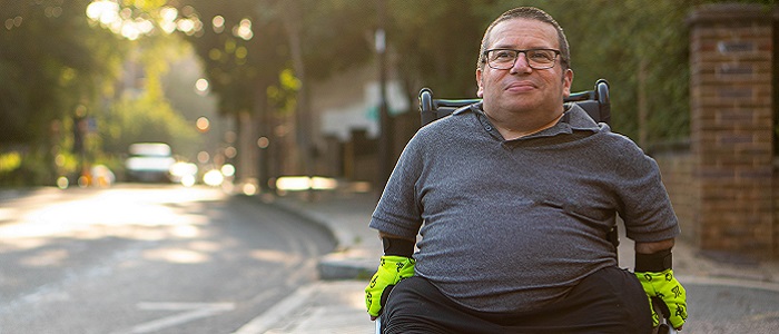 A young man in a wheelchair going along a surburban street in he sunshine. Source: Centre for Aging Better https://ageingbetter.resourcespace.com/pages/view.php?ref=10384&k=&search=age%2C+positive%2C+image%2C+library%2C+%40%40281&offset=240&order_by=resourceid&sort=DESC&archive=