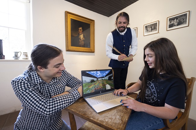In the kitchen of Robert Burns Ellisland Farm:  Left to right Bailey Hodgson, the University of Glasgow Minecraft Society’s President; Rhona Keenan, 11, from Dumfries using the Minecraft Ellisland game while been looked on by Richie Keenan, her father, dressed as Robert Burns, with a portrait of the poet in the background. Photo Credit Martin Shields