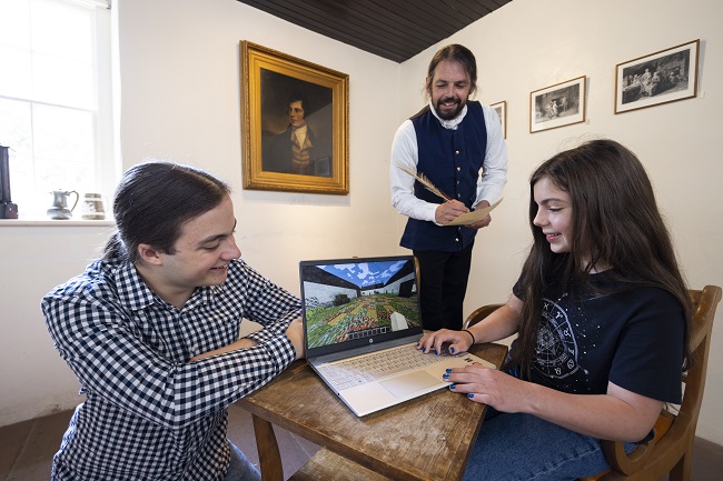 In the kitchen of Robert Burns Ellisland Farm: Left to right Bailey Hodgson, the University of Glasgow Minecraft Society’s President; Rhona Keenan, 11, from Dumfries using the Minecraft Ellisland game while been looked on by Richie Keenan, her father, dressed as Robert Burns, with a portrait of the poet in the background. Photo Credit Martin Shields