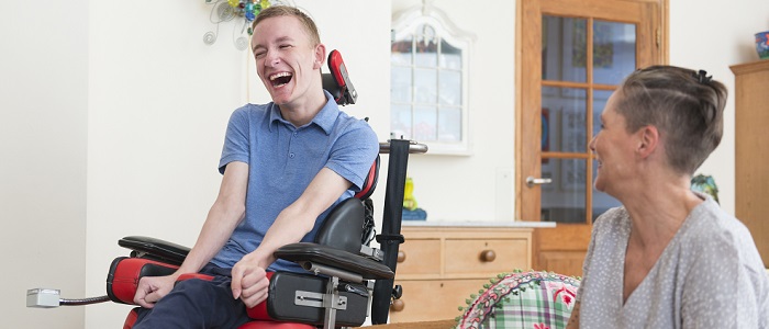 A young man with Amyotrophic lateral sclerosis in a wheelchair in a domestic lounge; he is laughing and sharing a joke with his mpother who is seated on a sofa next to him. Source: iStockPhoto https://www.istockphoto.com/photo/happy-young-als-patient-with-his-mom-gm603871162-103683657