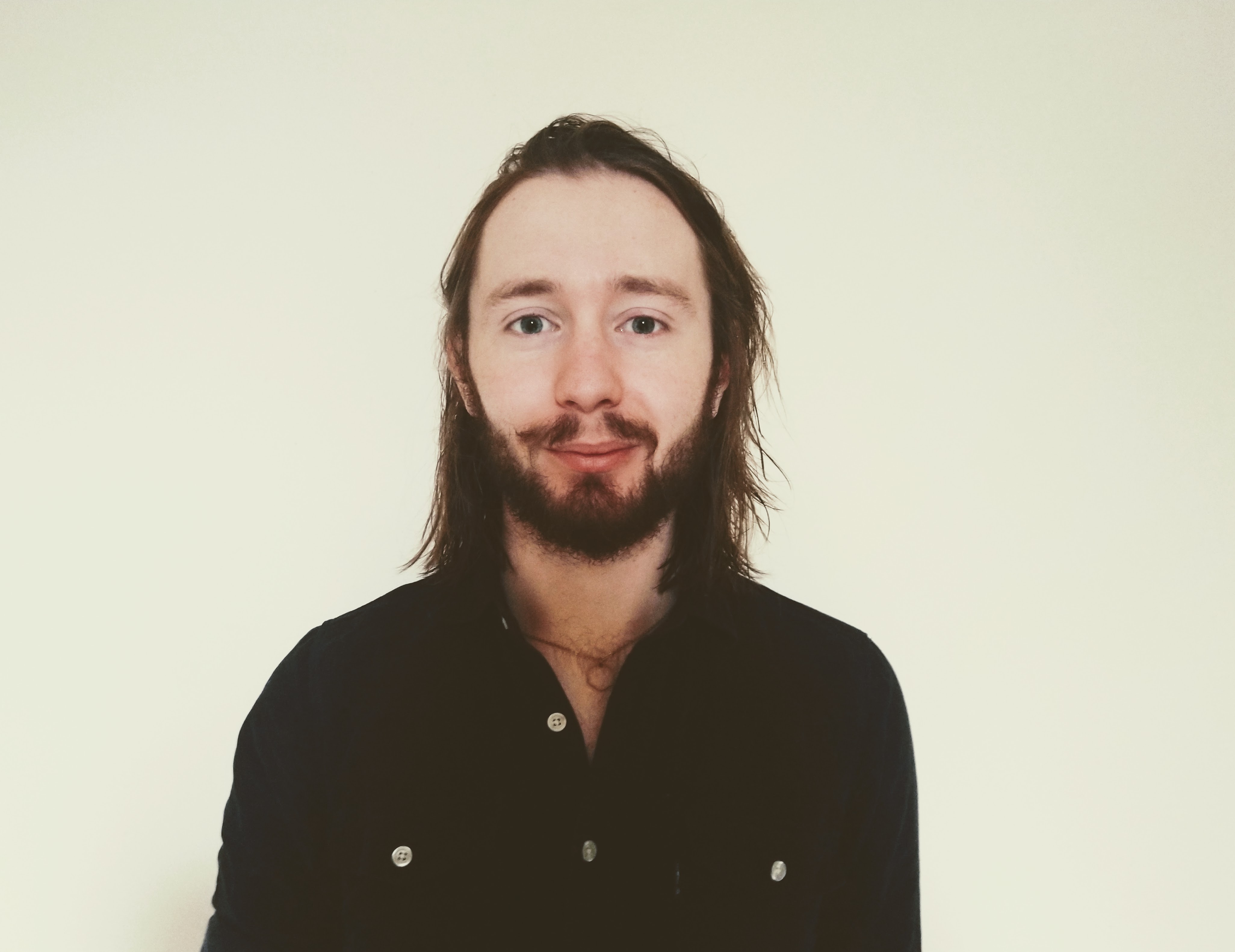 A passport style photo of Edward Curley. Image is at chest height showing a man wearing dark navy shirt, with shoulder length brown hair and similarly coloured beard. 